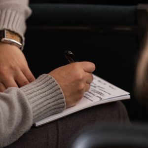 Person in grey sweater taking notes in a notebook with a pen.